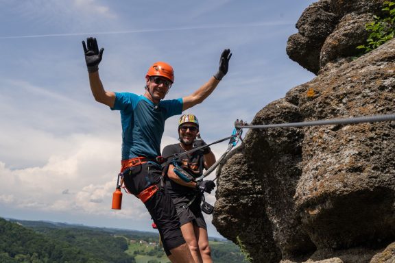 Zwei Kletterer an einem Felsen, einer winkt, der andere lächelt. Schönes Wetter und Landschaft im Hintergrund.