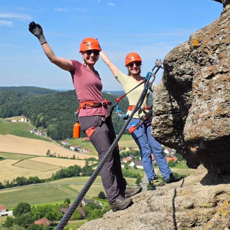 Zwei Personen in Kletterausrüstung lachen und posieren auf einem Felsen vor einer Landschaft.