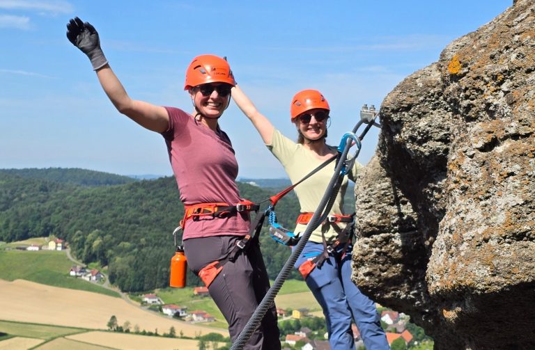 Leopold Klettersteig Zwei kletternde Personen auf einem Felsen, beide mit Helmen und fröhlichen Gesten.