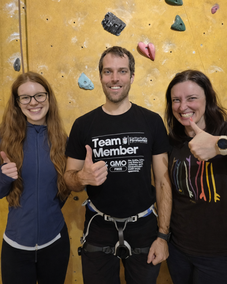 Drei Personen posieren an einer Kletterwand, zeigen Daumen hoch und lächeln.