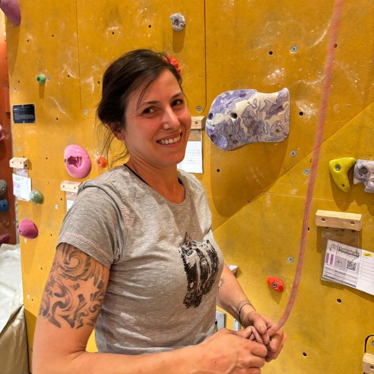 Frau mit tätowiertem Arm, die an einer Kletterwand eine Sicherung vorbereitet.