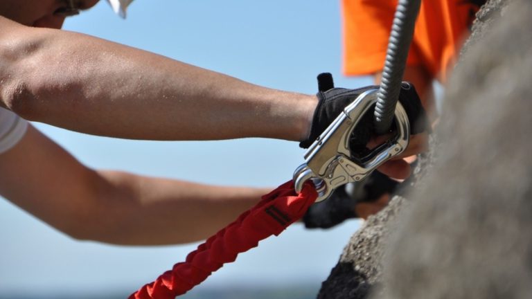Hände sichern einen Klettergurt an einem Felsen mit einem Karabiner.