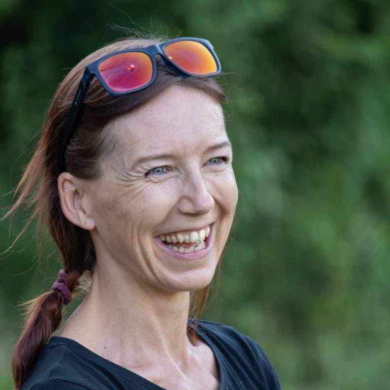 Nina Otopal Lächelnde Frau mit dunklem Haar in grauem Sweatshirt vor hellem Hintergrund.