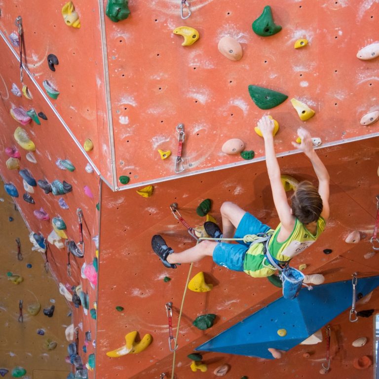 Junge klettert an einer farbigen Kletterwand in einer Halle.