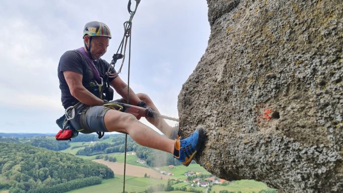 Kletterer, der an einer Steilwand hängt, mit Blick auf die Landschaft im Hintergrund.