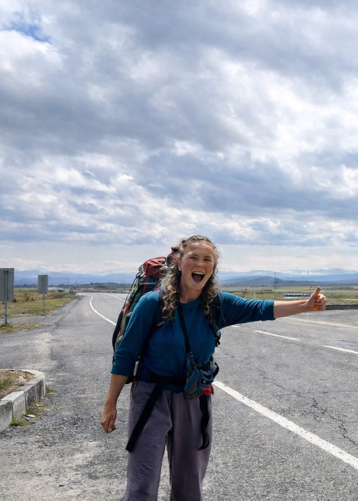 Lisa Janson Frau mit Rucksack steht an einer Straße und gibt Daumen hoch.